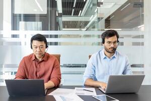 Two colleagues in casual clothes working inside office with laptops and headset. Office workers consulting clients remotely while sitting at desk. photo
