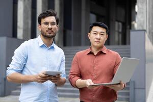 Two professional individuals standing outdoors, engaged in a business discussion with technology. The setting indicates a modern office environment, emphasizing collaboration and communication skills photo