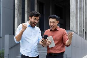 Two joyful colleagues cheerfully react to good news using a smartphone outdoors. Their jubilant expressions convey teamwork, achievement, and happiness in a professional setting photo