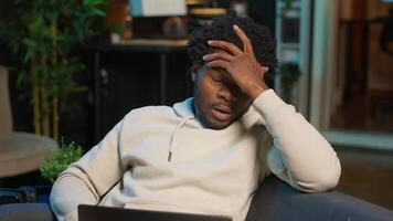 Self employed man suffering from pain and high fever sitting on sofa, trying to work and finish his tasks. Black male feelancer suffering from illness and feeling sick, using laptop. Camera B. video