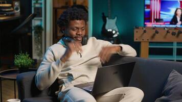 Freelancer listening to a podcast and being focused on online work, putting on headphones while seated on the couch in his living room. Productivity in a casual and flexible home. Camera A. video