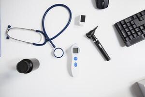 Top view shot of desk cluttered with stethoscope medication bottle thermometer mouse and keyboard symbolizing busy life of healthcare professional. Medical tools placed on doctors white table. photo