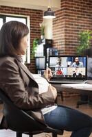 Selective focus of laptop displaying group conference call as pregnant businesswoman listens to her coworkers. Expectant lady with hand on pregnancy belly having videocall on digital device at office. photo