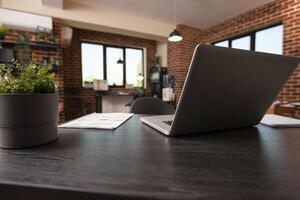 Closeup of desk arranged with digital device, documents and pot of plant in brick wall workspace. Vacant startup office creates professional look by having laptop and paperwork readily set on table. photo