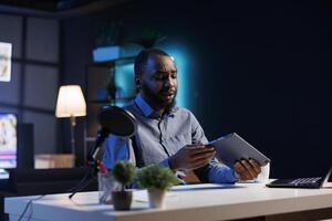 African american content creator analyzing tablet received from sponsor, doing honest review for online streaming channel. Internet show host reviewing digital device brand photo