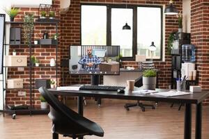 Vacant brick wall workplace setup with computer screen showing videocall of businessman in wheelchair. Desktop pc displaying ongoing virtual meeting with caucasian man in empty startup office. photo