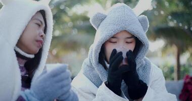 Handheld selective focus shot, Two Asian sibling dressed in warm clothes sit talking on chairs and enjoy a hot drink in the cold morning atmosphere. video