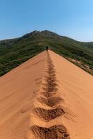 A lone figure walks along a sandy ridge towards a green mountain under a clear sky photo