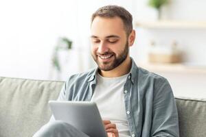 Joyful millennial man sitting on sofa with tablet computer, working or studying online at home. Happy Caucasian male participating in web lecture, taking part in webinar or conference photo