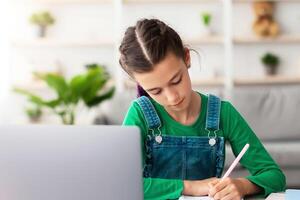 Front portrait of focused girl sitting at table with laptop computer in living room at home, writing essay or making test exam, taking notes in textbook, doing homework, free copy space photo