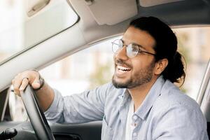 Joyful long-haired indian guy with glasses taxi diver driving brand new car, looking at rear view mirror and smiling, side view, copy space. Cheerful middle-eastern young man enjoying his new auto photo