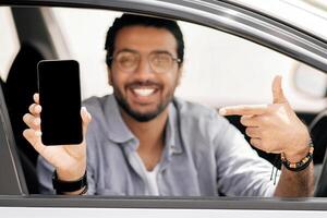 Selective focus on mobile phone with empty display in smiling middle-eastern guy hand, arab man driver using mobile application for navigation or car renting service, copy space, mockup photo