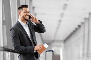 Mobile Call. Smiling Young Middle Eastern Businessman Talking On Cellphone In Airport While Waiting With Suitcase For Flight At Terminal, Happy Arab Man Having Pleasant Phone Conversation photo