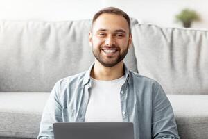 Full length of happy young guy sitting cross legged on floor, using laptop computer, smiling at camera indoors. Positive millennial freelancer working online from home, attending business webinar photo