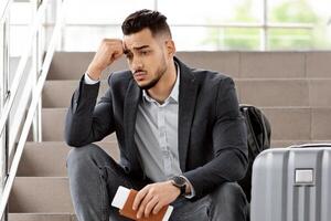 Upset Middle Eastern Guy Sitting On Airport Terminal Stairs With Luggage And Passport In Hands, Depressed Young Arab Man Missed His Flight, Touching Forehead And Thinking About Problem Solution photo