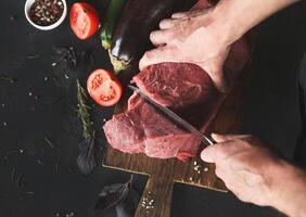 Man slicing filet mignon on wooden board at restaurant kitchen. Chef preparing fresh meat for cooking. Modern cuisine backgroung with herbs and vegetables, copy space, top view photo