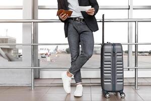 Unrecognizable Man Checking Boarding Time In Tickets While Standing With Luggage Near Window At Airport Terminal, Male Traveller Waiting For Flight, Ready For Trip, Cropped Image, Free Space photo
