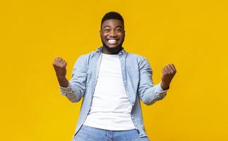 Portrait of overjoyed black man celebrating success with clenched fists and euphoric face expression on yellow studio background with copy space photo