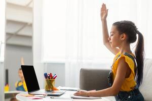 Homeschooling Concept. Profile side view of focused pupil sitting at desk on sofa, raising hand for an answer during call with teacher having virtual lesson, blank empty screen photo