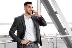 Mobile Communication. Handsome Middle Eastern Man Talking On Cellphone In Airport, Cheerful Young Arab Man Enjoying Pleasant Phone Conversation While Waiting For Departure In Terminal, Free Space photo