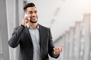Handsome Arab Entrepreneur Talking On Mobile Phone While Waiting For Flight Departure At Airport, Smiling Middle Eastern Businessman In Suit Standing At Terminal And Enjoying Cellphone Conversation photo