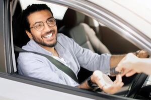 Cheerful young arab man sitting inside nice auto and taking automatic car key from leasing or renting service person, seller. Smiling indian guy doing test-drive before buying new automobile photo