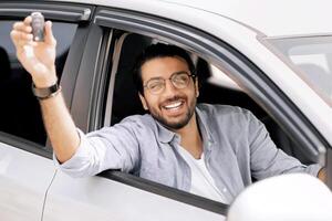 Cheerful indian long-haired guy with glasses sitting inside nice car and showing automatic key at camera, positive arab man renting automobile for travelling, having car ride or test-drive photo