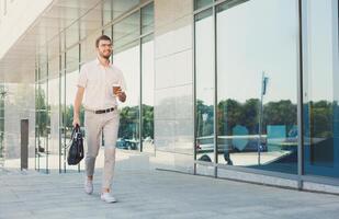 Young smiling businessman carrying briefcase and coffee cup while moving forward. Man in formalwear walking near modern office building, full length, copy space. Business and success concept photo