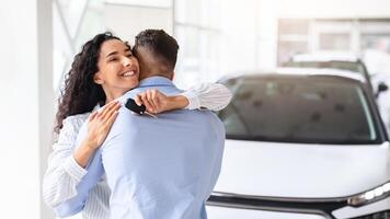 Ecstatic young curly woman with key from brand new car hugging her loving husband and smiling, feeling thankful and happy, standing by luxury auto, showroom interior, panorama with copy space photo