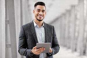 Smiling Young Middle Eastern Man With Digital Tablet In Hands Posing At Airport Terminal, Successful Millennial Arab Businessman Using Tab Computer While Waiting For Flight Boarding, Copy Space photo