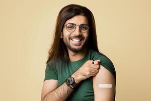Handsome millennial long-haired indian man got vaccinated against COVID-19, showing shoulder with plaster on and smiling at camera, sitting on chair over beige studio background, copy space photo