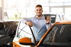 Wealthy middle-eastern businessman buying new car in luxury auto showroom, cheerful arab guy standing by nice comfy yellow automobile, showing key and smiling at camera, copy space photo
