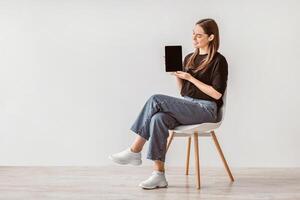 Charming young woman showing tablet with empty screen, sitting on chair against white studio wall, mockup for website or new app design. Millennial lady demonstrating touch pad display template photo