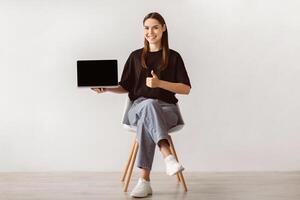 Smiling young woman sitting on chair, showing laptop with empty screen, gesturing thumb up, mockup for website. Millennial lady with portable computer recommending your product ad photo