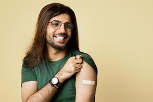 Happy millennial arab guy got vaccinated against COVID-19, showing shoulder with plaster on and smiling, sitting on chair over beige studio background, looking at copy space, panorama photo