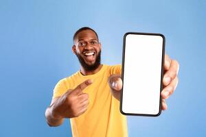 Mobile advertisement. Overjoyed African American man pointing at cellphone with empty white screen on blue studio background, mockup for app or website. Cellphone display template photo
