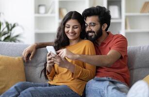 Portrait Of Happy Indian Couple Resting With Smartphone On Couch, Smiling Eastern Spouses Shopping Online Or Browsing New Mobile App While Relaxing In Living Room, Closeup Shot With Free photo