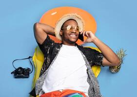 Funky black guy in trendy summer clothes lying on inflatable lilo, speaking on smartphone over blue studio background, top view. African American man having phone conversation on beach vacation photo