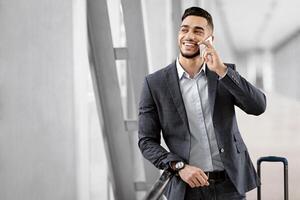 Handsome Young Arab Businessman Talking On Cellphone While Waiting In Airport Terminal, Young Middle Eastern Man Standing Near Boarding Gate, Enjoying Pleasant Mobile Phone Conversation, Free Space photo