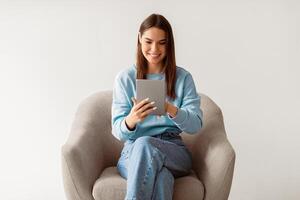 Young Caucasian woman using digital tablet, browsing internet or social media, sitting in armchair, enjoying modern technologies, checking new app or website on white studio background photo
