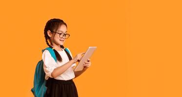 E-Learning. Asian School Girl Using Digital Tablet Doing Homework Online Standing Over Yellow Studio Background. Panorama With Empty Space photo