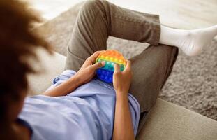 Unrecognizable black teenager playing with POP IT toy, using antistress sensory plaything, sitting on sofa at home, closeup. Cropped view of African American youth enjoying relaxing game photo