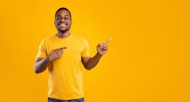 Look There. Cheerful African American Man Pointing Fingers Aside Showing Empty Space For Text Posing Smiling To Camera Over Yellow Background In Studio. Panorama photo