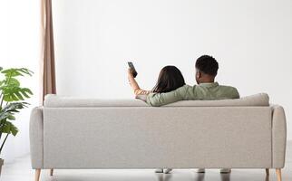 Back view of hugging black lovers sitting on couch at home, watching TV, unrecognizable african american man aiming remote controller towards copy space on white wall, panorama, full length photo