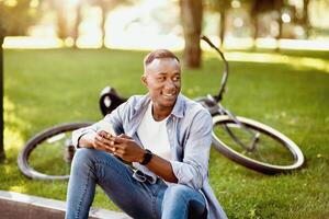 Cool black guy with modern bike using smartphone at park. Positive African American man with mobile device checking messages, reading book, watching or browsing internet outside photo