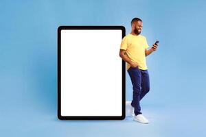 Optimistic young African American man leaning on huge tablet computer with blank white screen, using mobile phone on blue studio background, mockup for website or application design photo