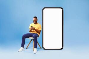 Cheerful young African American man sitting on chair near big smartphone with empty screen, using mobile device, presenting space for website, advertisement or mobile app, blue studio background photo