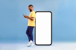 Full length of young African American guy using cellphone while leaning on giant mobile phone with empty white screen, blue studio background. Mockup for app or website, space for advertisement photo