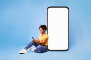 Cheery African American lady sitting near big cellphone with empty white screen, using mobile device, checking new cool app on blue studio background, mockup for website or application design photo