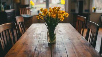 A wooden dining table is adorned with a mason jar holding vibrant yellow daffodils, bringing a cheerful touch to the room. Natural light fills the space, enhancing the spring atmosphere. photo